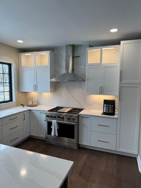 Kitchen range wall: a stainless gas range and chimney hood against a full-height white veined backsplash, with lit glass-front upper cabinets on both sides.