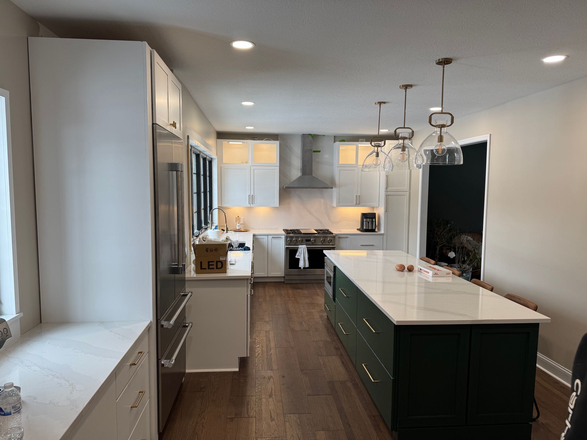 Kitchen with white shaker cabinets and a long island painted deep green, white veined countertops, three glass pendants, a stainless range under a chimney hood and wide-plank wood floors.