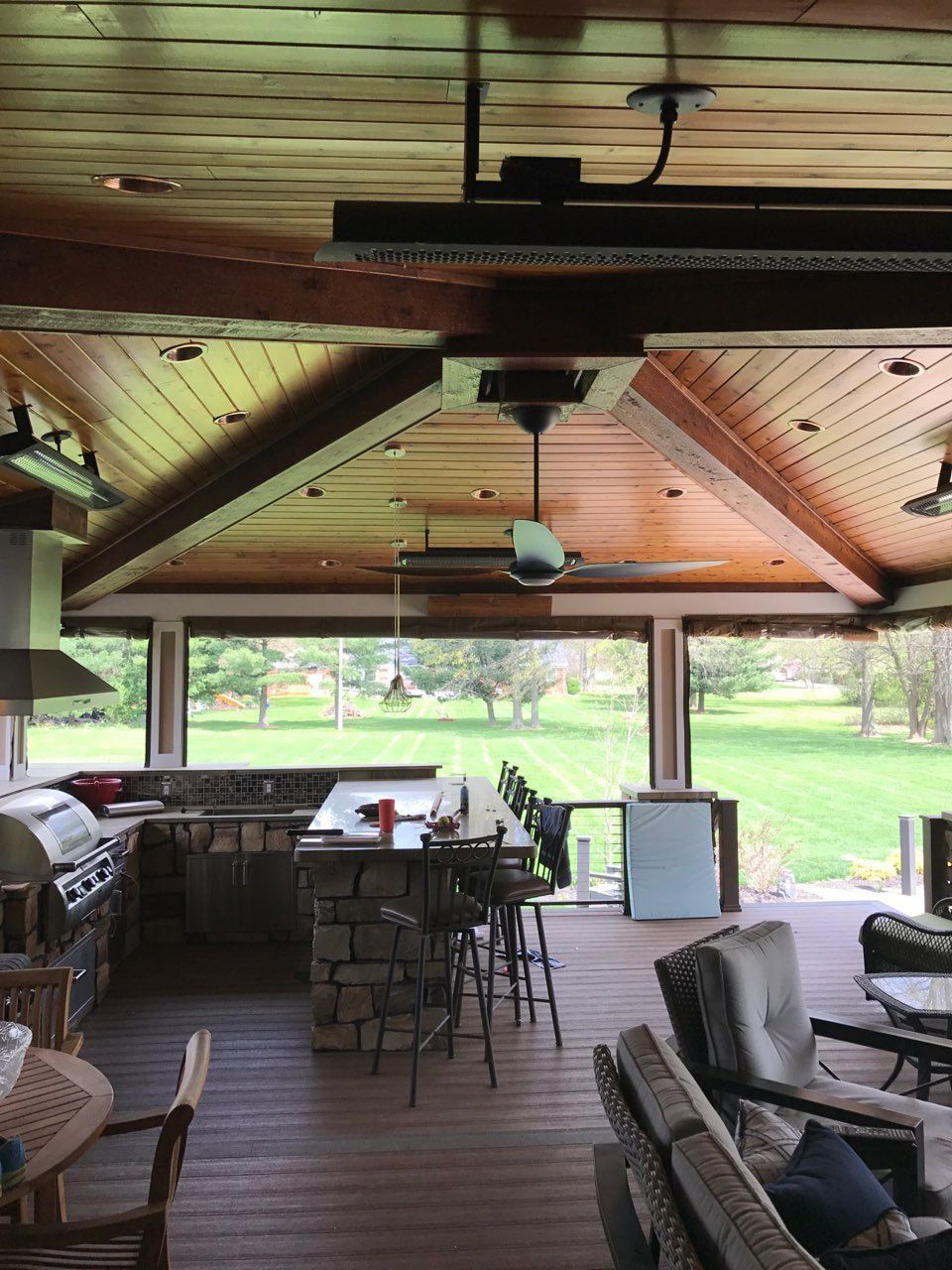 Outdoor kitchen pavilion: a wood plank ceiling with timber trusses, a ceiling fan and overhead heaters above a stone-faced bar with stools, on a deck facing a wide lawn.