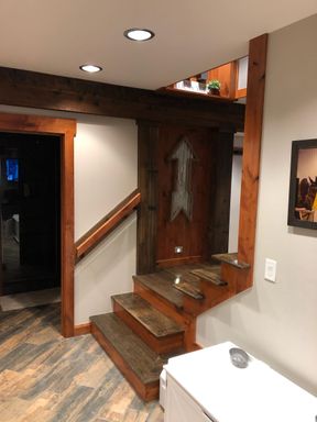 Basement stair: stained wood treads turning up past a dark plank wall, with wood trim, a wood handrail and a timber beam above.