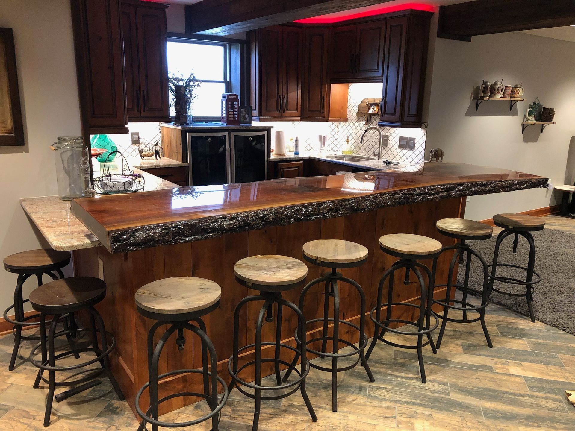 Basement bar with a live-edge wood top, dark cabinets and a tiled backsplash behind it, and a row of wood-seat stools on a plank floor.