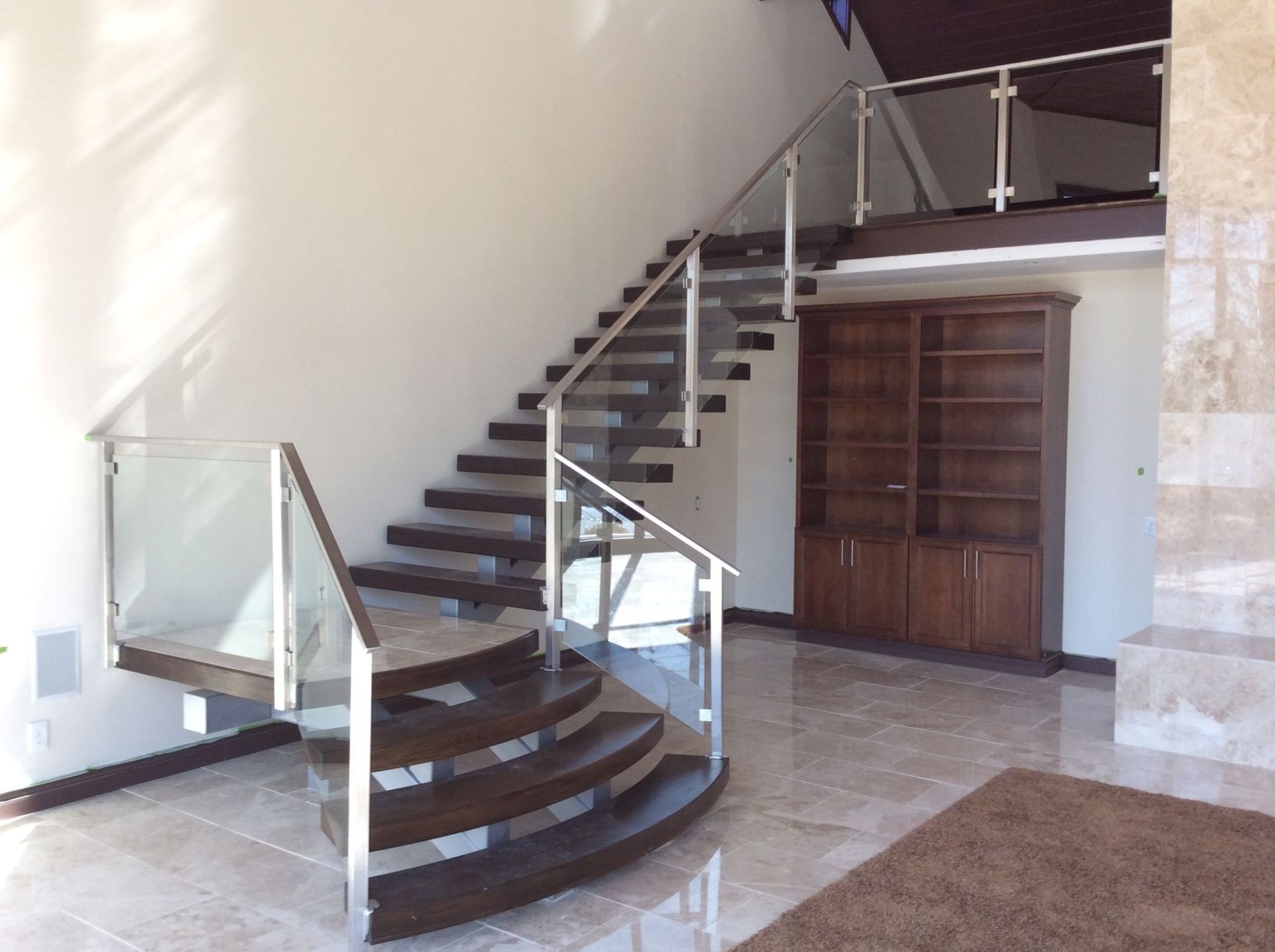 Entry hall: a curved open-tread staircase with dark wood treads and a glass panel railing on steel posts, rising from a polished stone floor past a wood built-in under the landing.