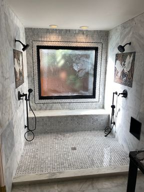 Bathroom walk-in shower for two: a window framed in mosaic tile between two black shower heads and hand showers, a tiled bench under the window, marble-look walls and a mosaic floor.
