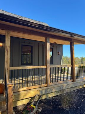 Covered porch from the yard: cedar posts and a cedar top rail with black balusters, under the roof edge of a board-and-batten house.