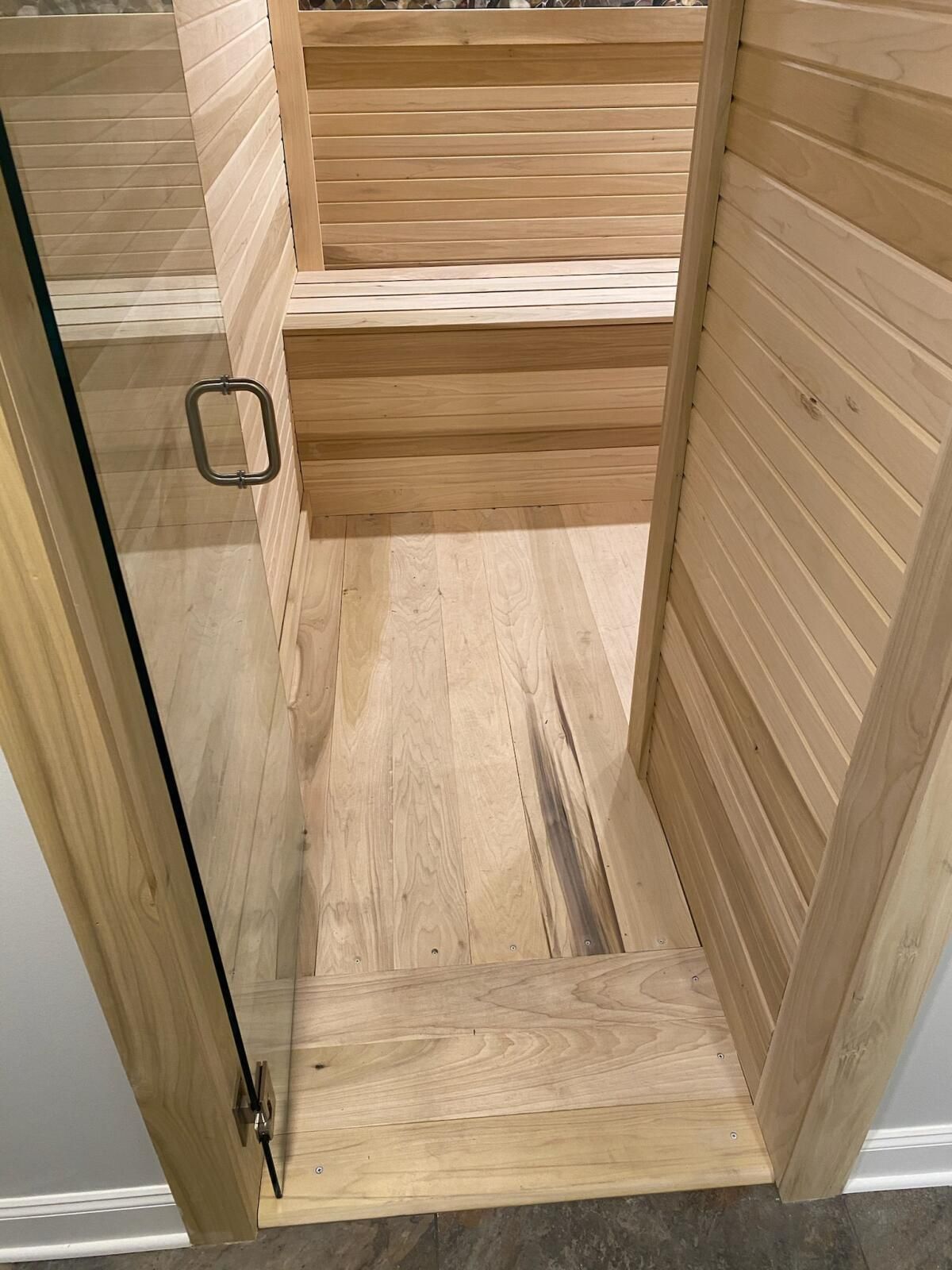 Home sauna: a glass door open onto a small room lined in light wood boards, with a slatted bench along the back wall and a wood board floor.