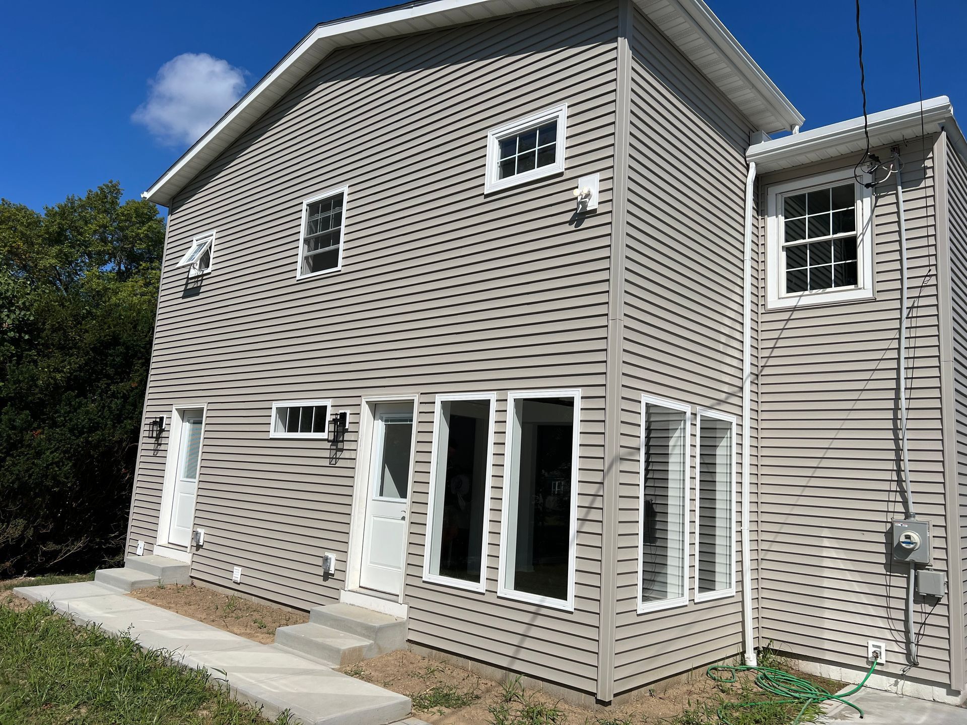 House exterior: a two-story addition in gray lap siding under its own gable roof, with new windows on both floors, two entry doors and a new concrete walk and steps.