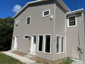 House exterior: a two-story addition in gray lap siding under its own gable roof, with new windows on both floors, two entry doors and a new concrete walk and steps.