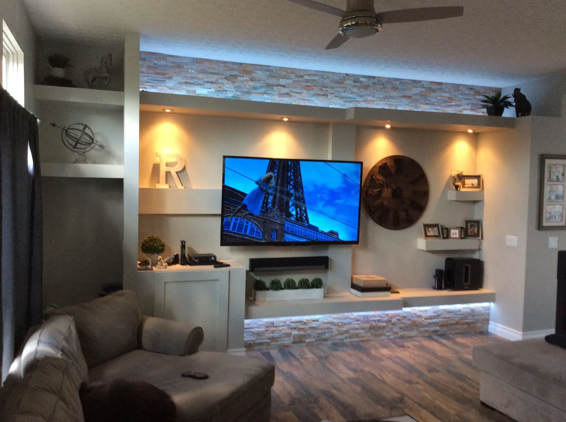 Basement media wall: a TV between lit niches and floating shelves, a stacked-stone ledge along the top, built-in cabinets below and a lit stone strip along the base.