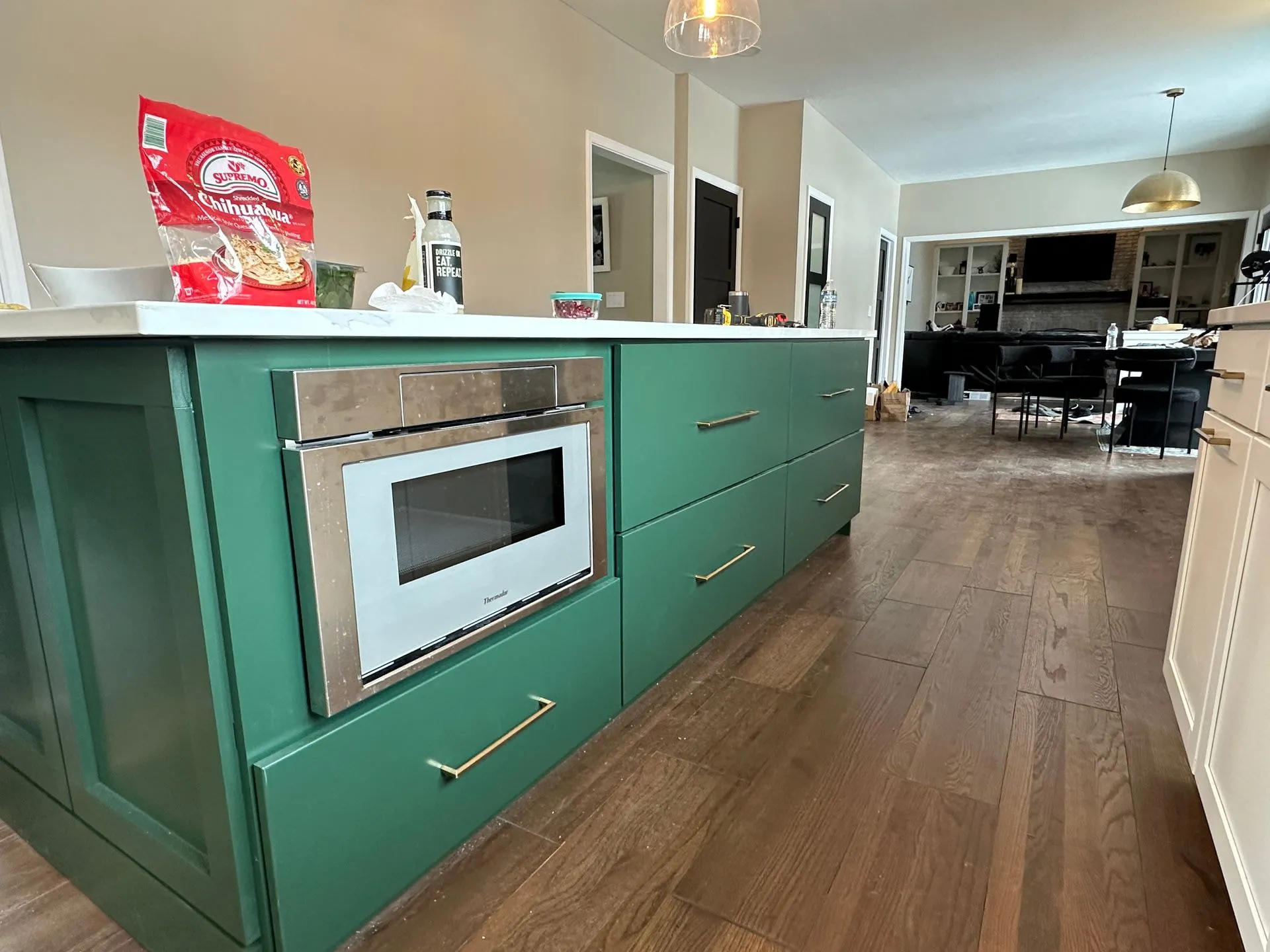 Kitchen island, working side: green shaker drawers with brass bar pulls and a stainless microwave drawer, with the wide-plank floor running toward the family room.