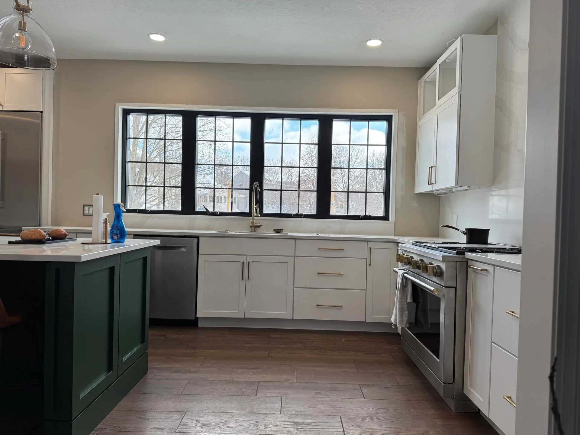 Kitchen sink wall: four black-framed windows over white shaker base cabinets and a dishwasher, with a stainless range at right and the end of the green island at left.