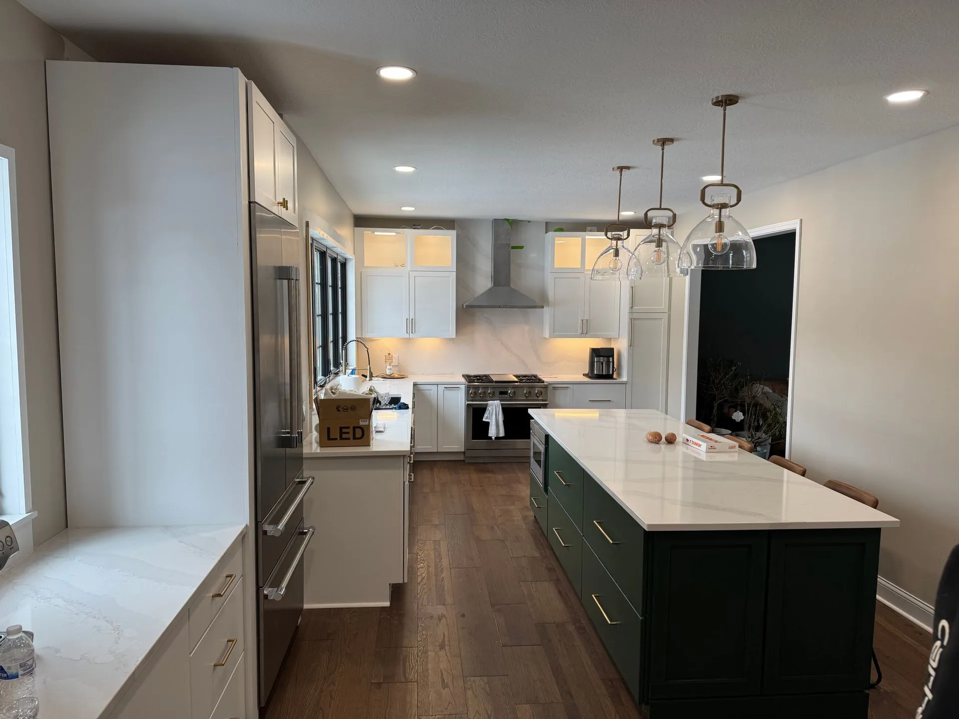 Kitchen with white shaker cabinets and a long island painted deep green, white veined countertops, three glass pendants, a stainless range under a chimney hood and wide-plank wood floors.
