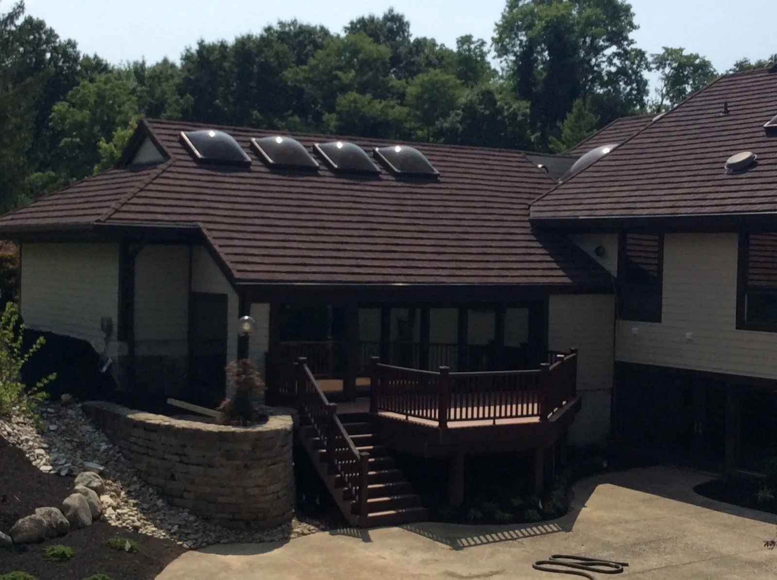 Back of a house: a raised deck with a stained railing and stairs down to a concrete patio, beside a curved stone retaining wall, under a long roof with dome skylights.