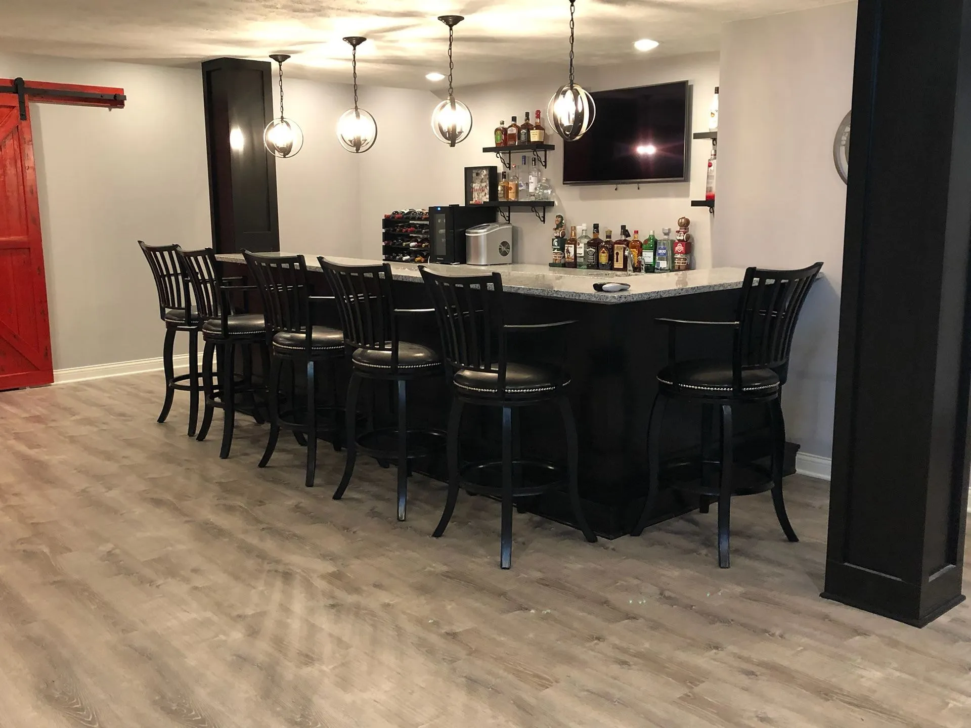 Basement bar: a long dark bar with a stone top and black stools, glass globe pendants, and bottle shelves and a TV on the wall behind.