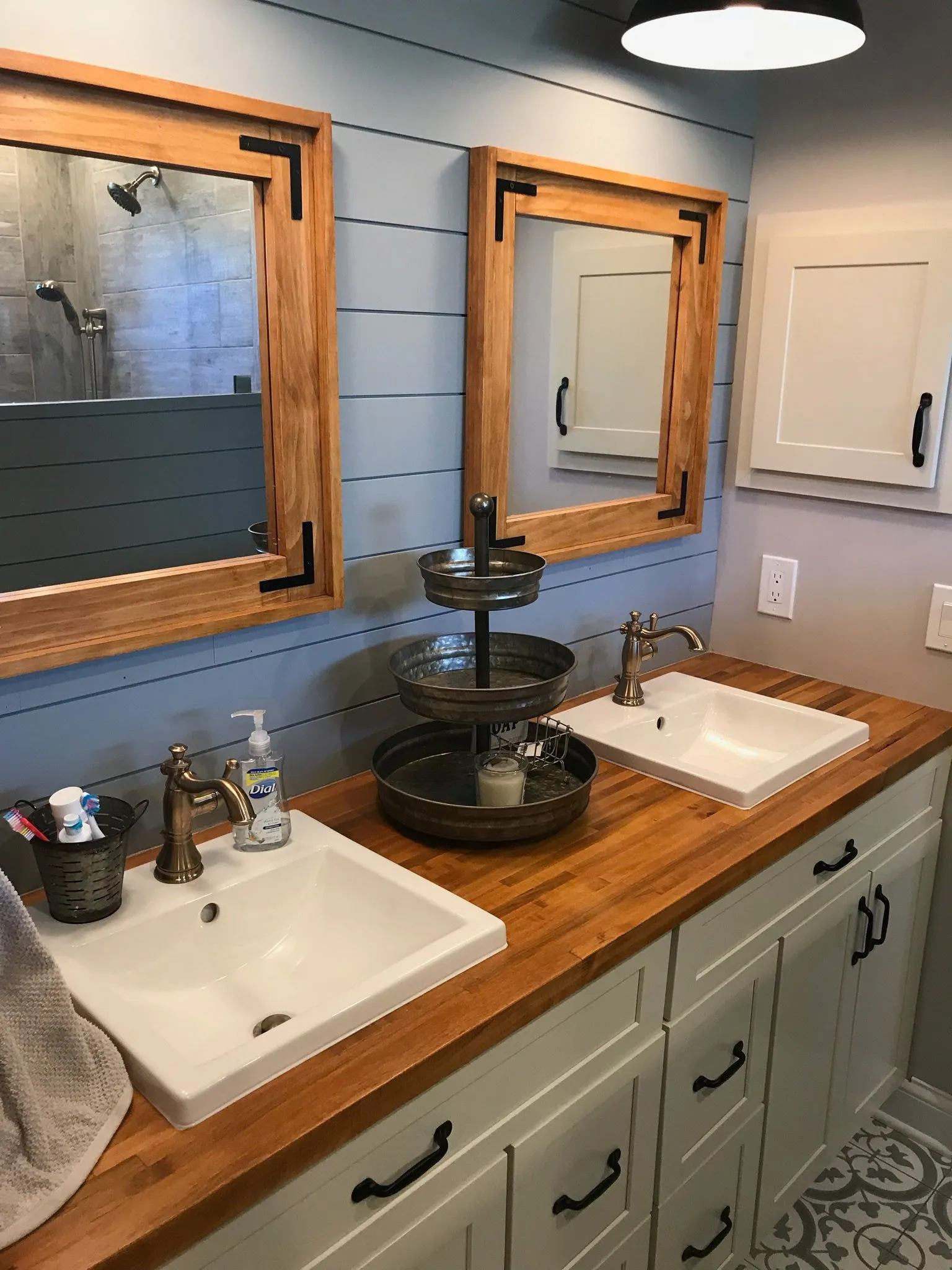 Bathroom: a double vanity with two white sinks set in a wood countertop, bronze faucets, wood-framed mirrors on gray-green shiplap, white cabinets with black pulls and a patterned tile floor.