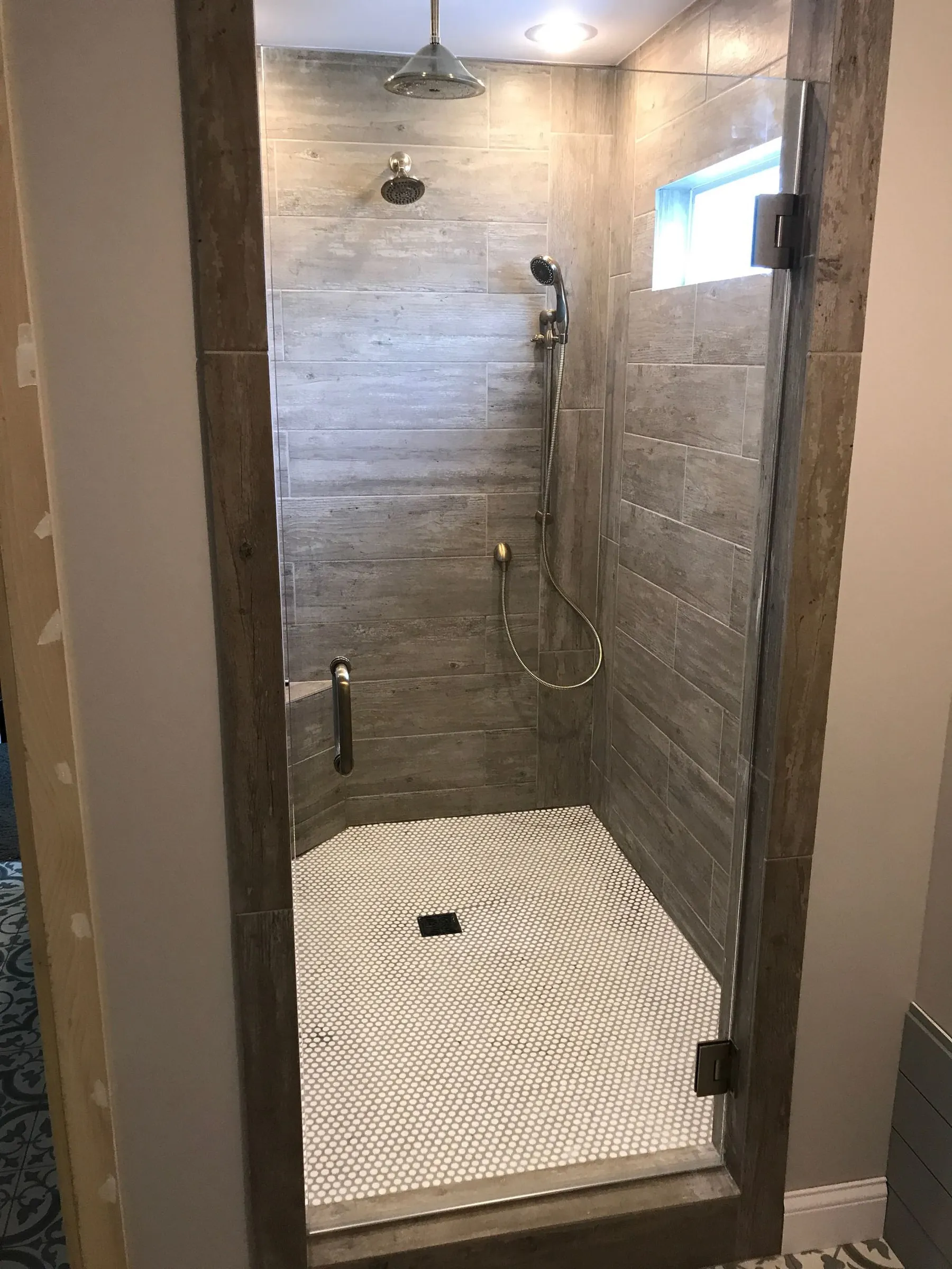 Bathroom shower: gray wood-look plank tile walls, a rain head and a hand shower, a grab bar, a white penny-tile floor and a frameless glass door.