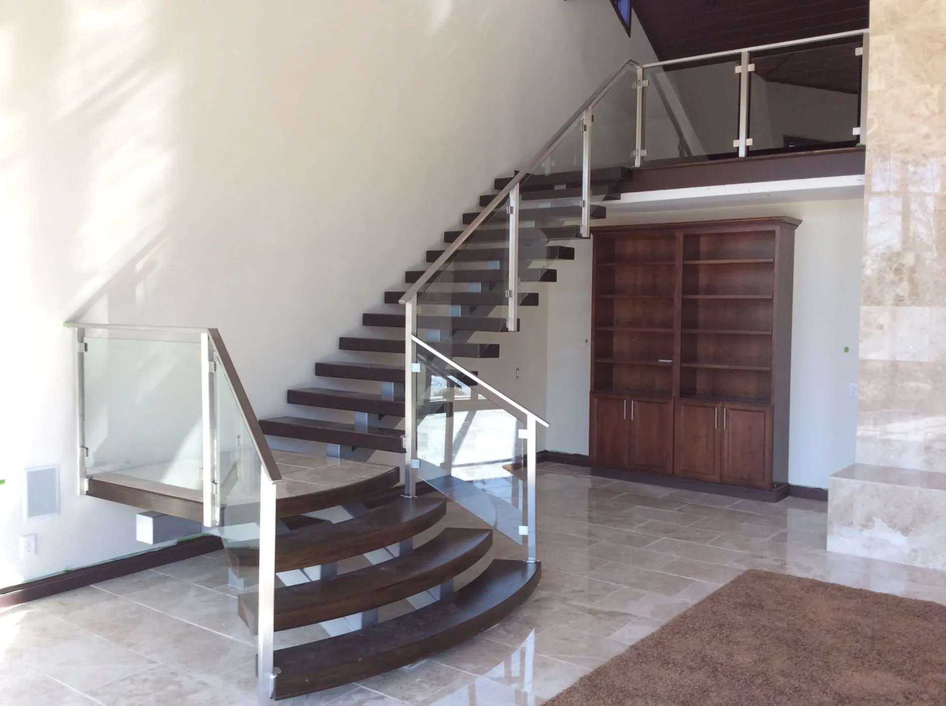 Entry hall: a curved open-tread staircase with dark wood treads and a glass panel railing on steel posts, rising from a polished stone floor past a wood built-in under the landing.