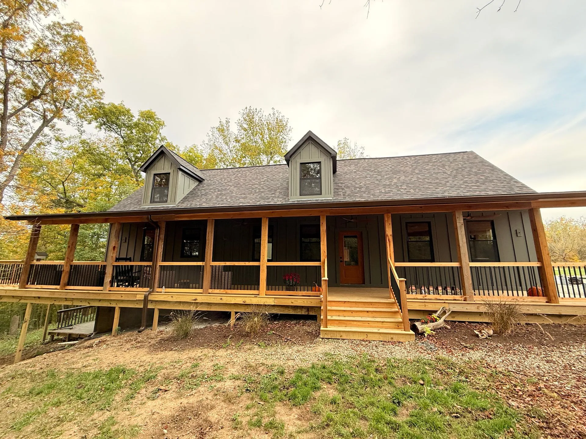 House exterior: a story-and-a-half house with two front dormers in board-and-batten siding, a long front porch on cedar posts with a black-baluster railing, and wide cedar steps.