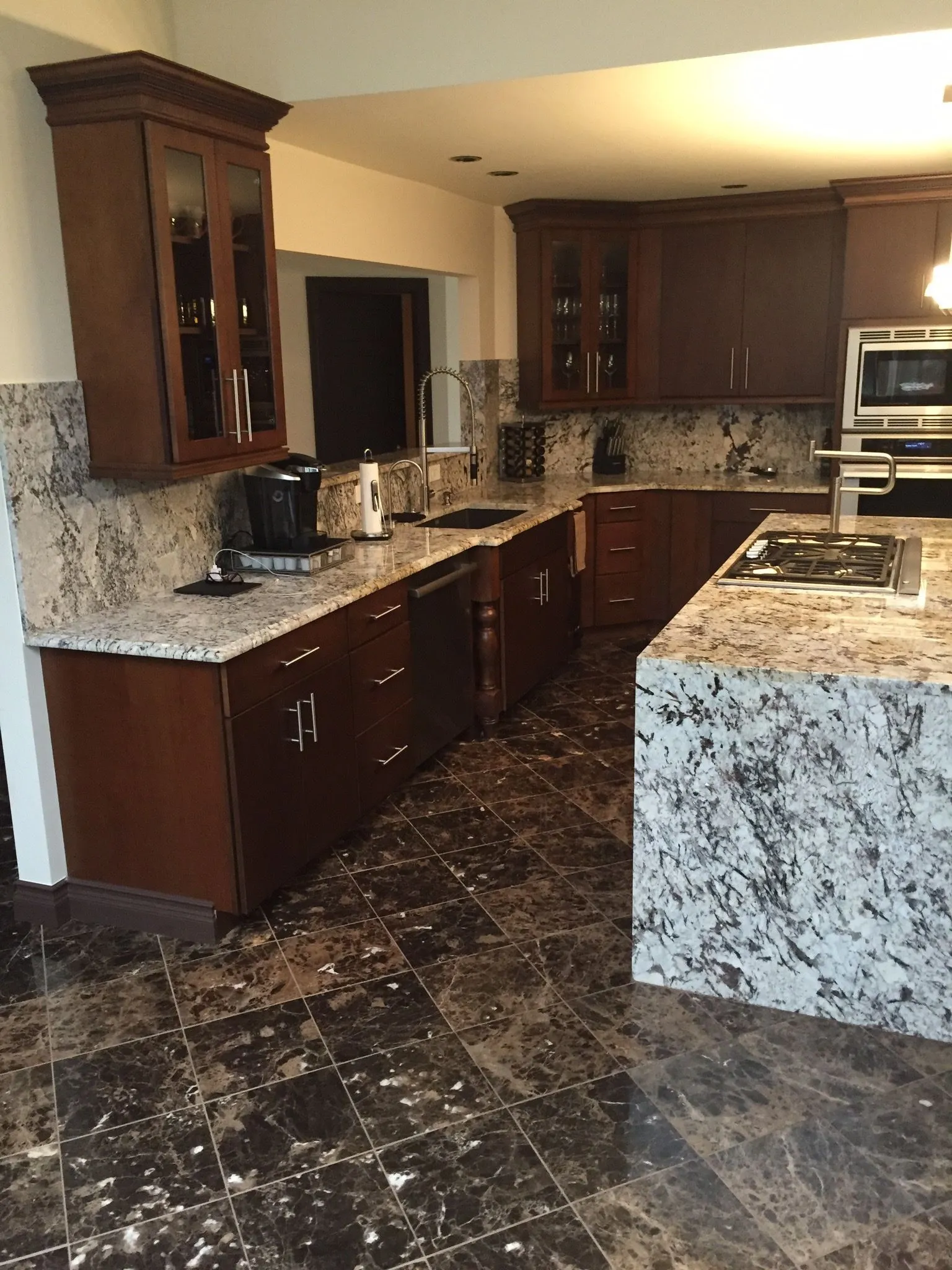 Kitchen with dark wood cabinets and glass-front uppers, granite counters and backsplash, a granite waterfall island with a gas cooktop, and a polished dark stone floor.