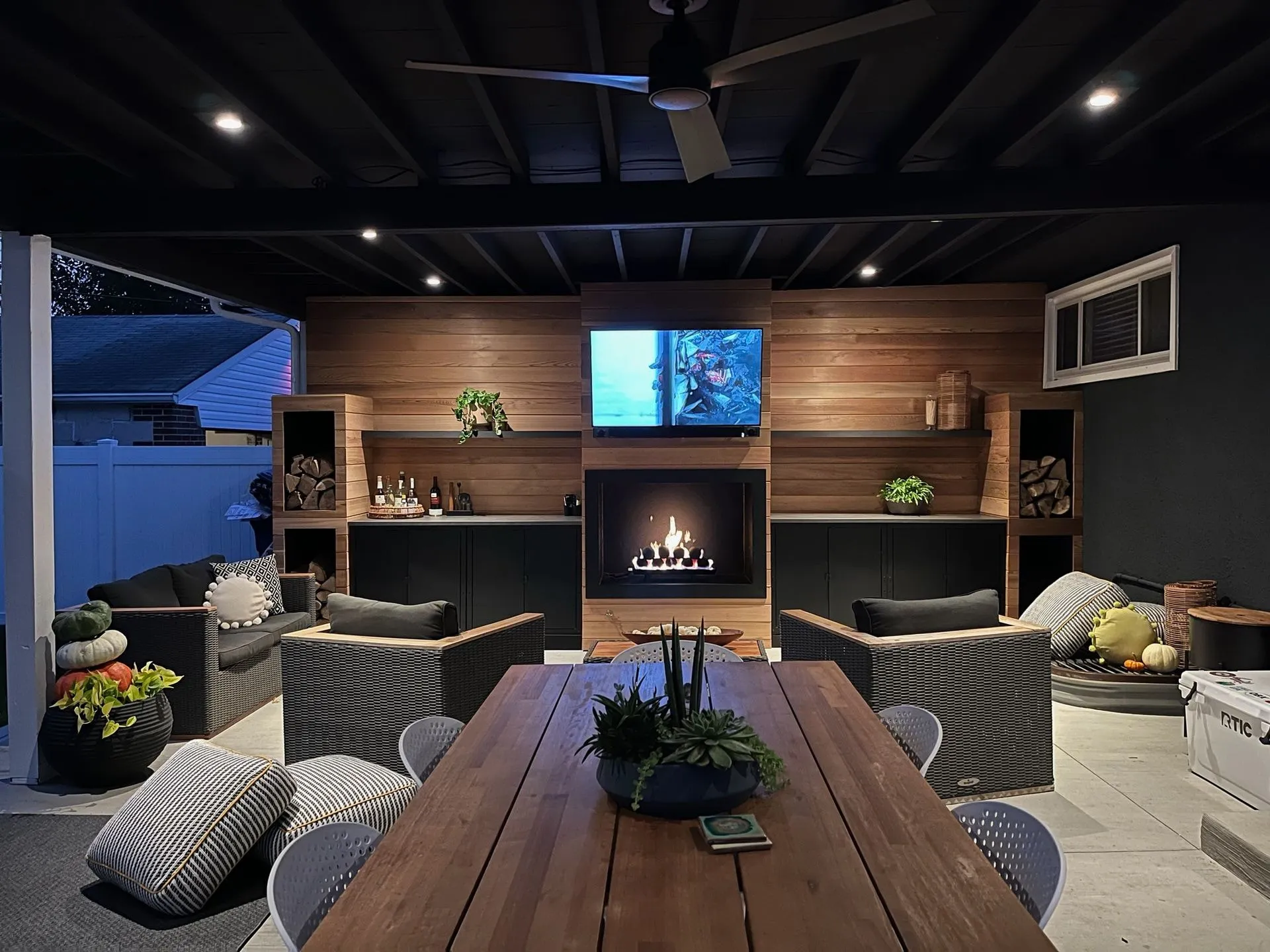 Covered patio at dusk: a cedar plank wall with a lit fireplace and a TV above it, log cubbies and shelves on both sides, recessed lights in a dark ceiling and a long wood dining table in front.