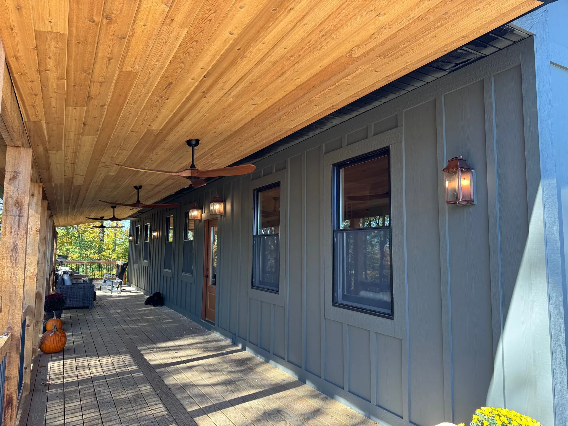 Covered porch: a tongue-and-groove cedar ceiling with ceiling fans, copper lanterns on a board-and-batten wall, black-framed windows and a wood deck floor running the length of the house.