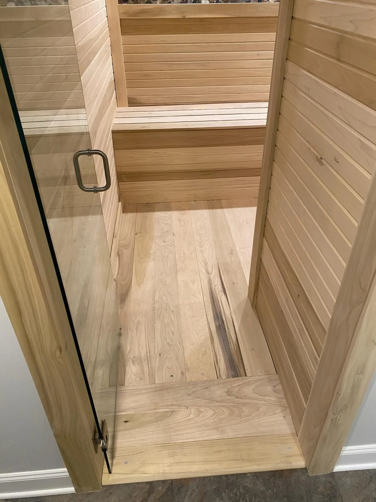 Home sauna: a glass door open onto a small room lined in light wood boards, with a slatted bench along the back wall and a wood board floor.